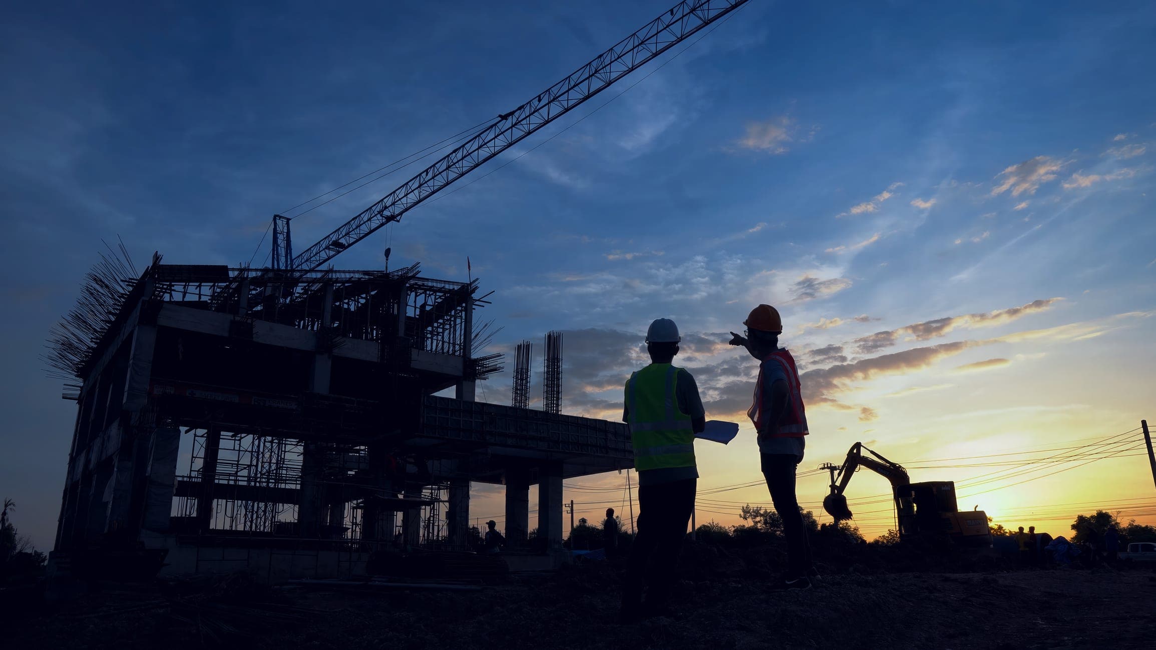 Two site leads in hard hats reviewing drawings at dusk beside a building under construction